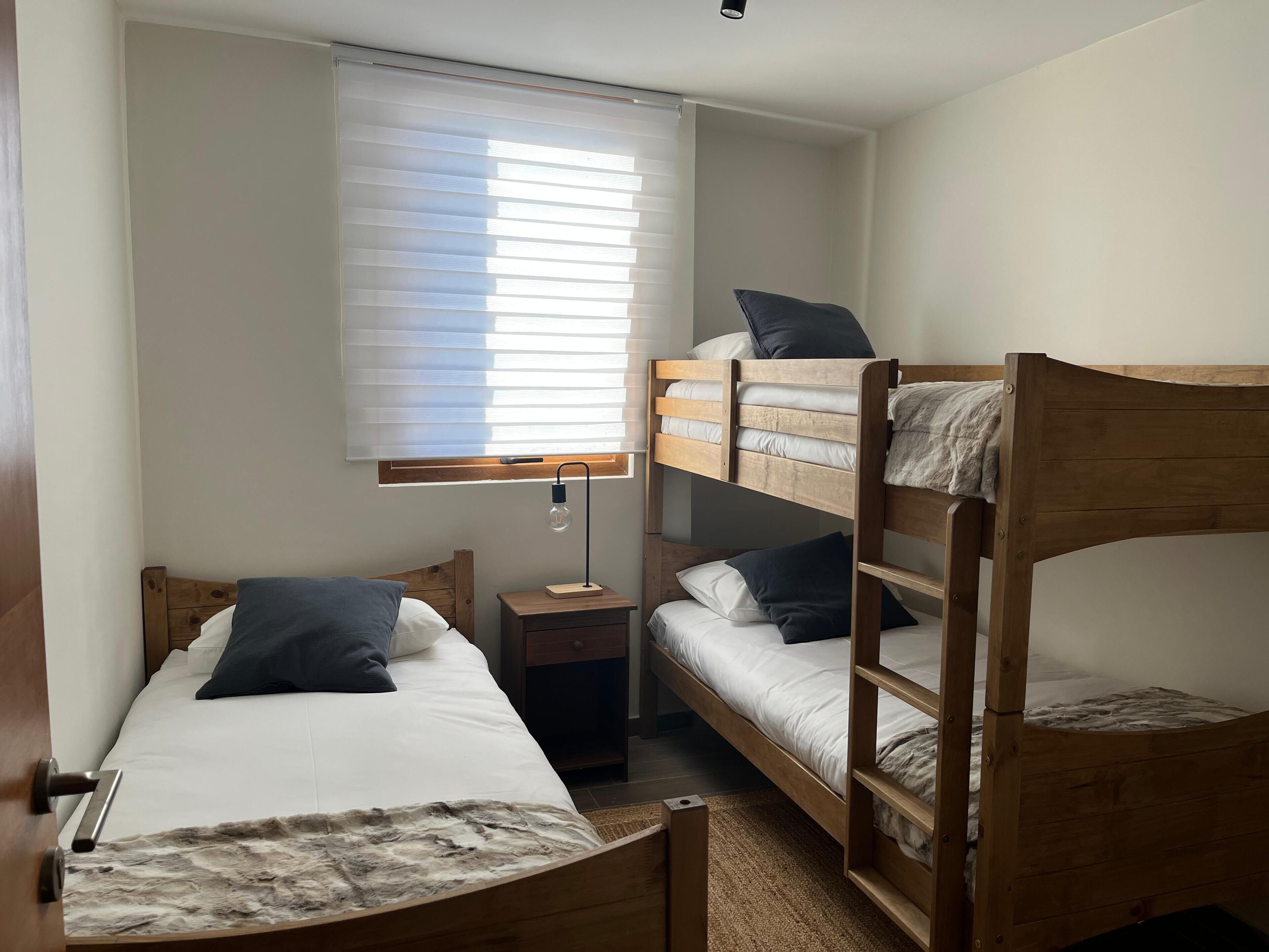 A bunk bed room with a window, a bedside table, and a lamp.