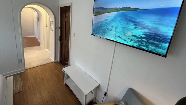 Una habitación con un televisor de pantalla plana grande montado en la pared, piso de madera, un banco blanco y una puerta que da a otra habitación.