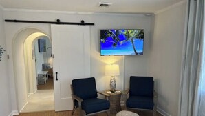 Una habitación con dos sillas azules, un televisor en la pared, una lámpara y un otoman pequeño y redondo.