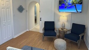 Una sala de estar con dos sillones azul oscuro, un televisor montado en la pared, una lámpara sobre una mesita auxiliar y un otoman estampado.