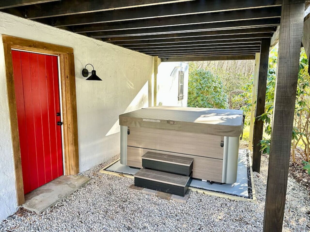 A hot tub under a wooden pergola with a red door and a wall-mounted light.