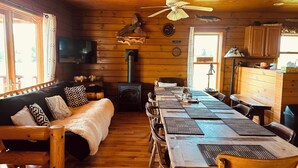 A cabin interior with a wooden floor, a sofa, a dining table with chairs, a fireplace, a ceiling fan, and a television mounted on the wall. - Twin Chalets of Finger Lakes (Geneva)