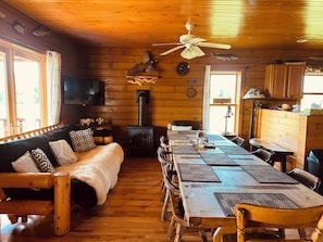 L’intérieur d’une cabane est doté d’un plancher en bois, d’un canapé, d’une table à manger avec des chaises, d’une cheminée, d’un ventilateur de plafond et d’un téléviseur fixé au mur.