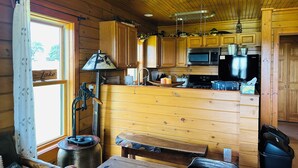 A cabin interior with wooden walls, a kitchen area with a microwave and stove, a dining table, and a window with curtains. - Twin Chalets of Finger Lakes (Geneva)