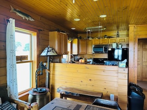 A cabin interior with wooden walls, a kitchen area with a microwave and stove, a dining table, and a window with curtains. - Twin Chalets of Finger Lakes (Geneva)