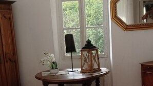 Ein Schlafzimmer mit einem Bett, einem runden Holztisch mit Lampe, einem Spiegel und einem Fenster mit Blick auf Grünflächen.