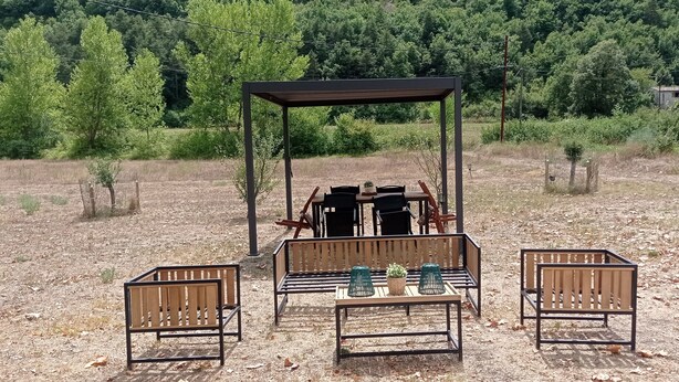 Ein Sitzbereich im Freien mit Tisch und Stühlen unter einer hölzernen Pergola, umgeben von einer natürlichen Landschaft.