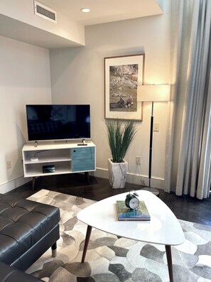 A modern living room with a flat-screen TV, a white coffee table with a book, a black leather sofa, a marble-patterned rug, a potted plant, and a framed picture on the wall.