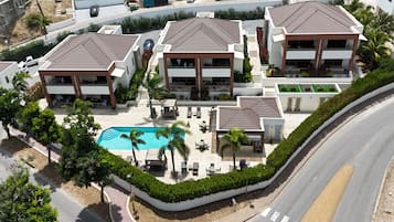 Een modern resort met meerdere gebouwen, een centraal zwembad en een aangelegde tuin met palmbomen en ligbedden.