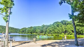 A lakeside beach with a sandy shore, a dock with boats, and a forested hill in the background.