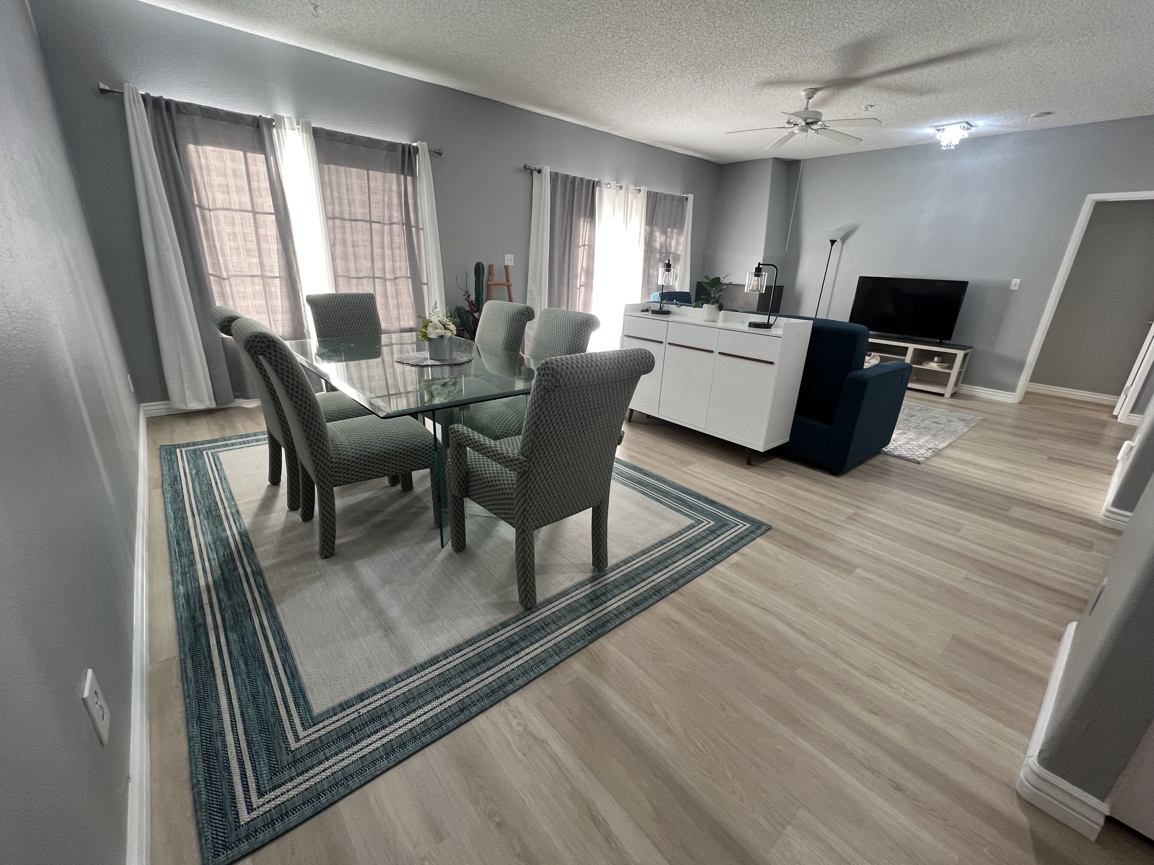 A modern living room with a glass dining table, four patterned chairs, a white cabinet, a flat-screen TV, and a ceiling fan.