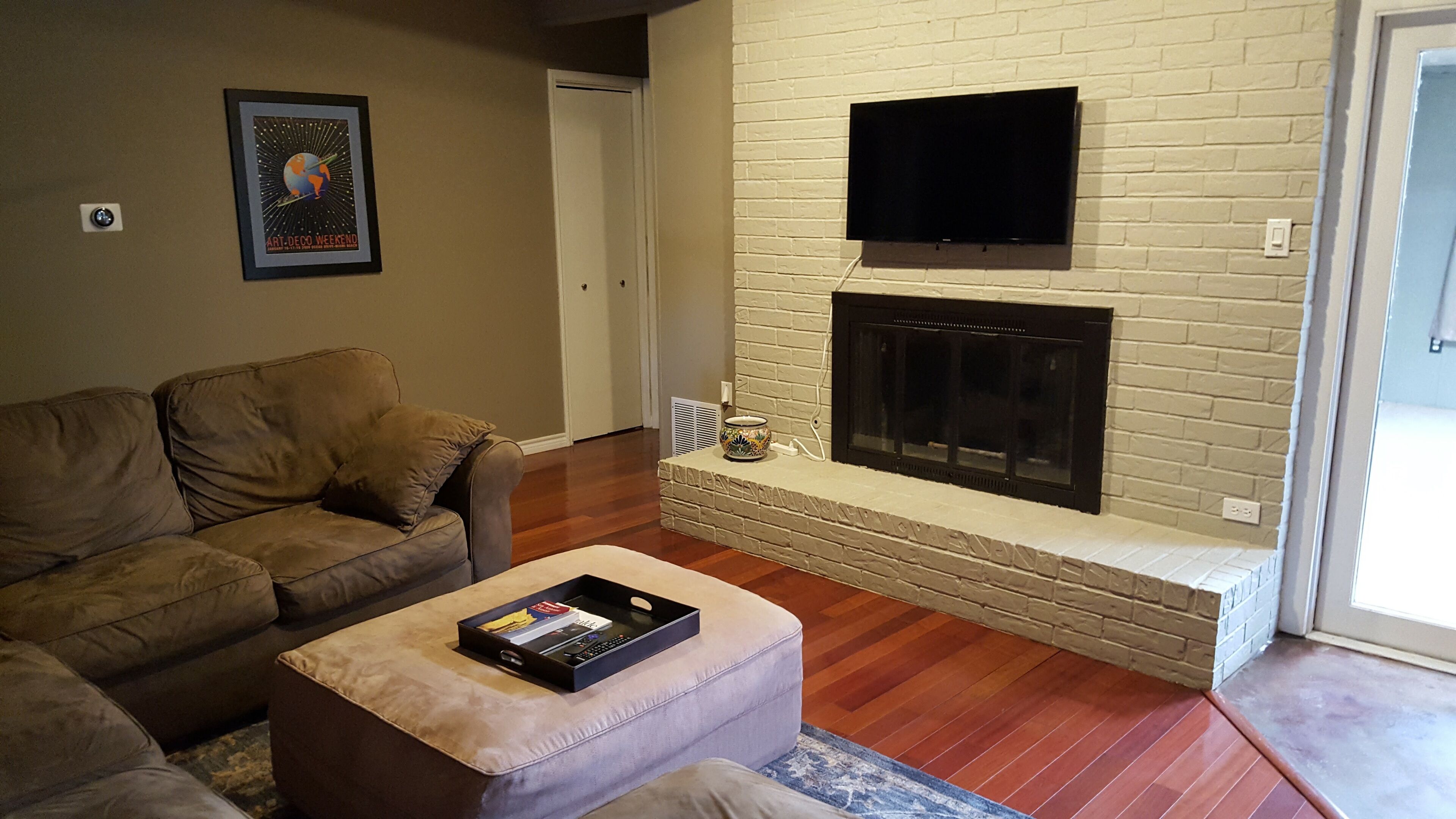 A living room with a beige sofa, a small ottoman with a tray, a fireplace, and a flat-screen TV mounted above it.