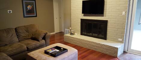 A living room with a beige sofa, a small ottoman with a tray, a fireplace, and a flat-screen TV mounted above it.