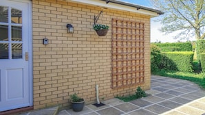 Un bâtiment en briques avec une porte blanche, une fenêtre, un luminaire fixé au mur, un pot de fleurs suspendu et un panneau en treillis de bois.