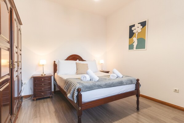 A neatly made bed with a wooden headboard, two bedside lamps, and a small wooden nightstand with drawers. - Avelino Apartment by Atlantic Holiday (Machico)