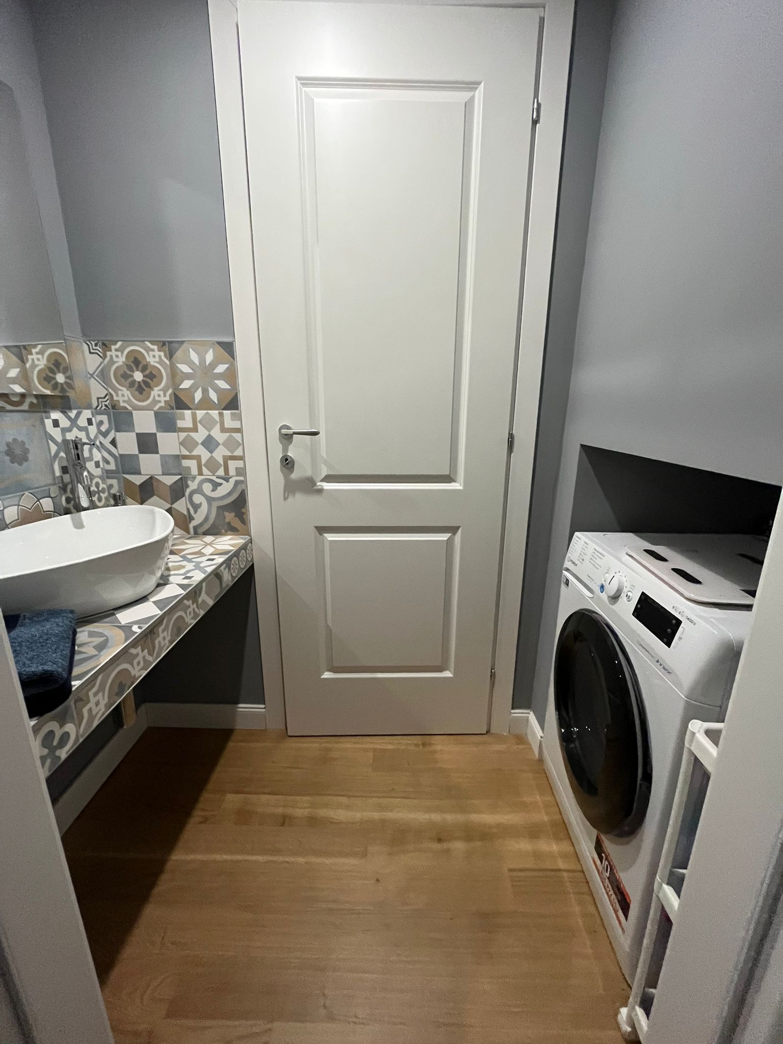 Een kleine wasruimte met een wasmachine, een wastafel en een wand betegeld met decoratieve tegels.
