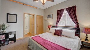 A bedroom with a bed, a ceiling fan, a window with curtains, a bedside lamp, a bookshelf, and a door. - Studio Condo Near Lake Pepin - King Bed - Dewdrop (Pepin)