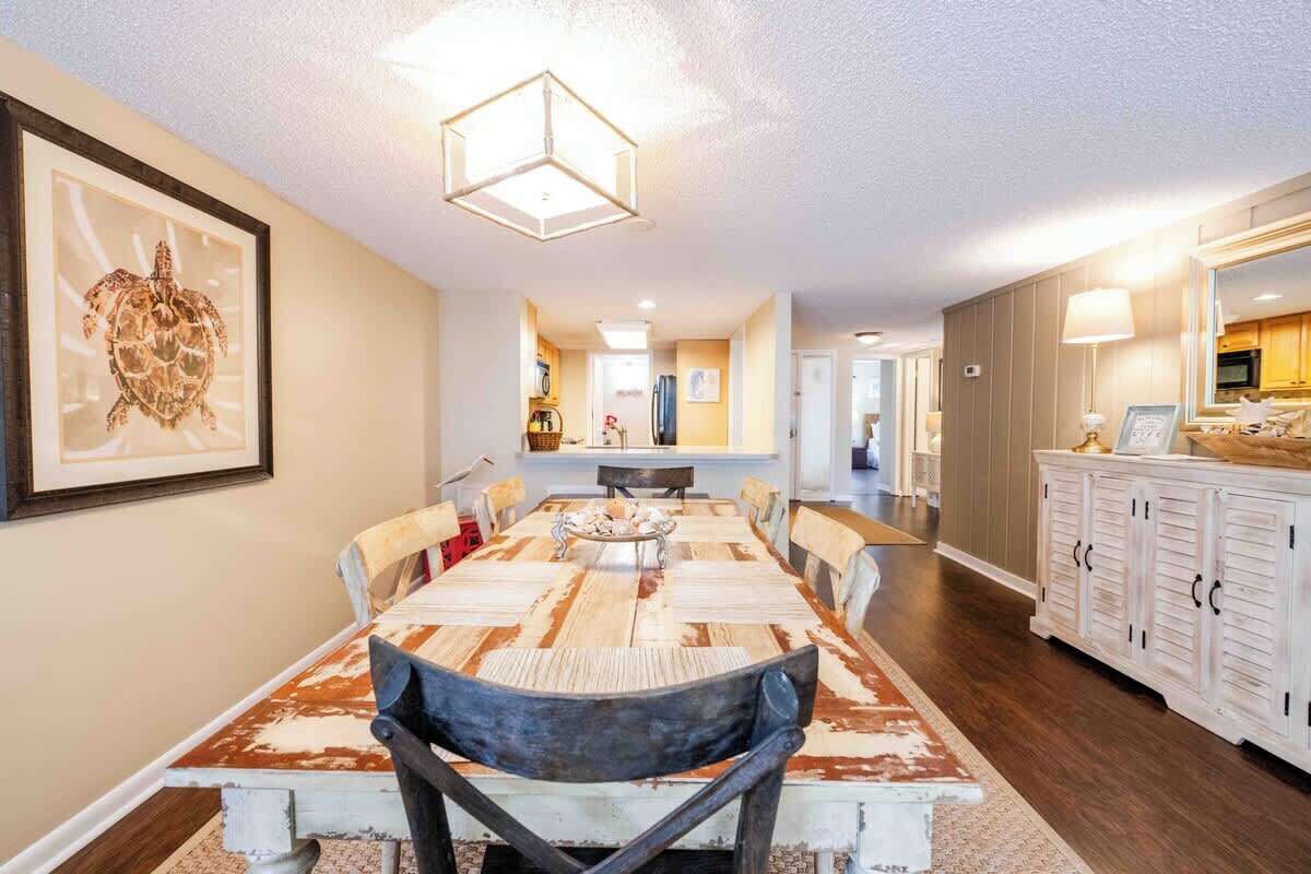 A dining area with a long wooden table, chairs, and a chandelier. There is a kitchen visible in the background.