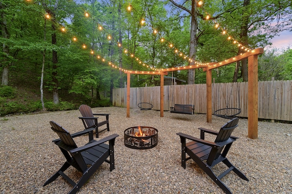 Un espacio al aire libre con asientos, una chimenea, sillas de madera y luces colgantes.