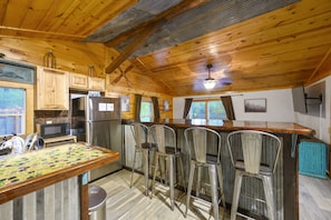 Cocina rústica con vigas de madera, refrigerador de acero inoxidable y una zona de bar con taburetes metálicos.