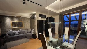 Ein modernes Wohnzimmer mit einem Glastisch, einem grauen Sofa und Blick auf die Stadt durch große Fenster.