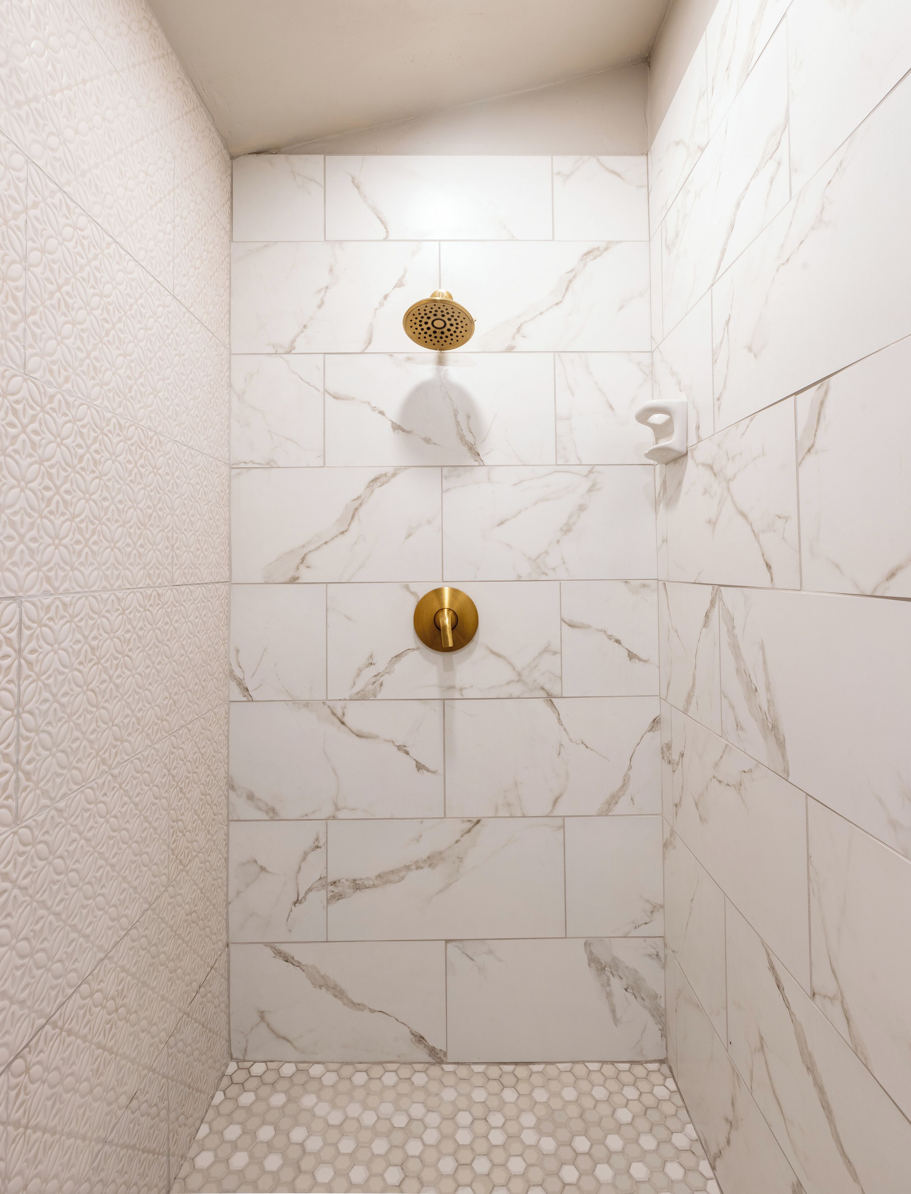 Une douche aux parois recouvertes de carreaux de marbre blanc et dotée d’un pommeau de douche doré.