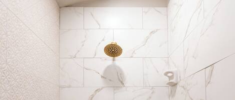 Une douche aux parois recouvertes de carreaux de marbre blanc et dotée d’un pommeau de douche doré.