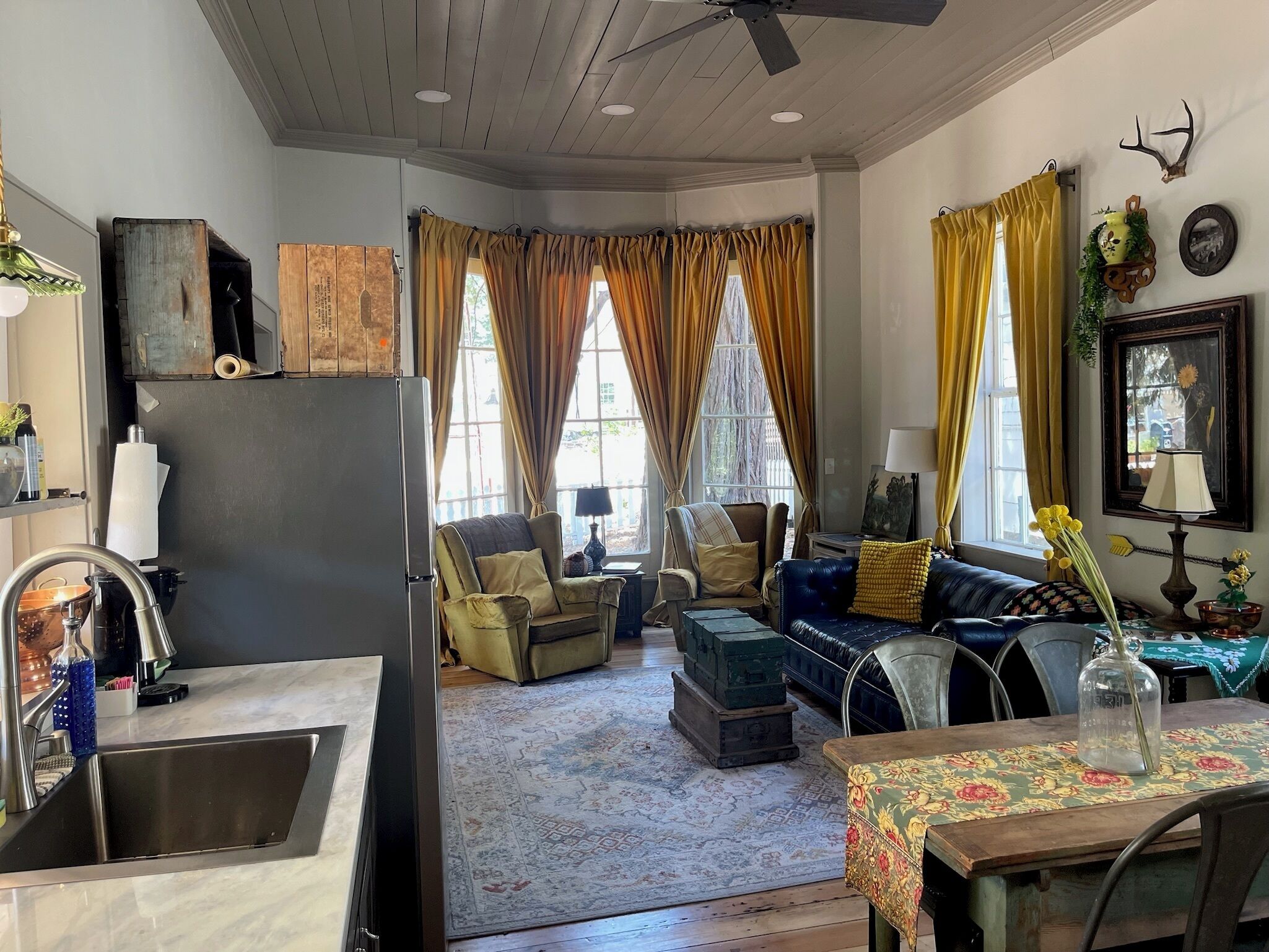A cozy living room with a sofa, armchairs, and a dining table. There is a kitchen area with a sink and a refrigerator.