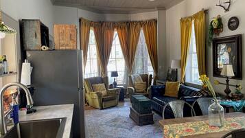 A cozy living room with a sofa, armchairs, and a dining table. There is a kitchen area with a sink and a refrigerator.
