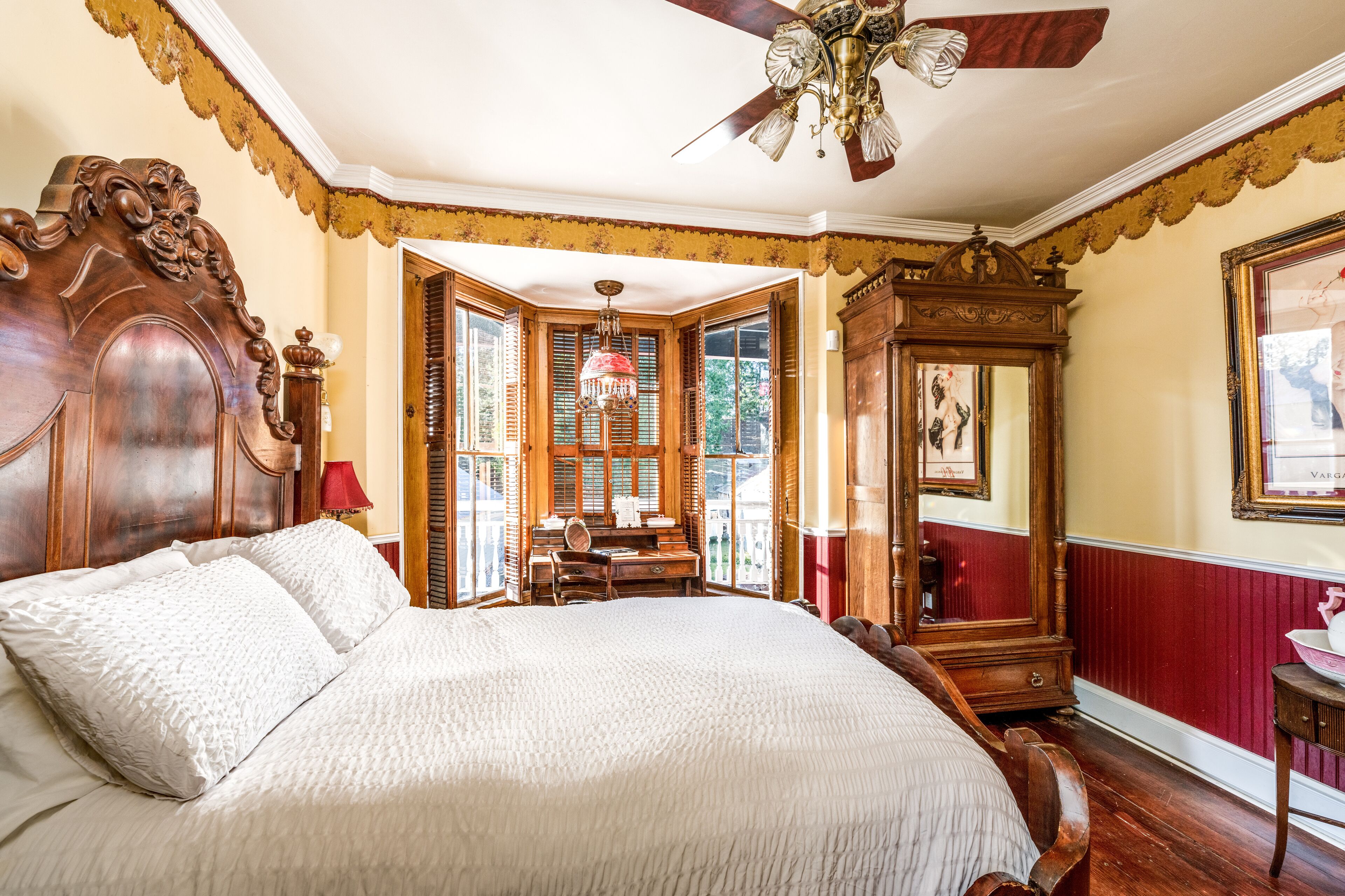 Un dormitorio con una cama grande, cabecera de madera, ventilador de techo, cómoda de madera y un espejo con marco rojo.