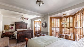 Un dormitorio con una cama grande, un aparador de madera, un escritorio y una silla. Hay una ventana con persianas de madera y una araña.