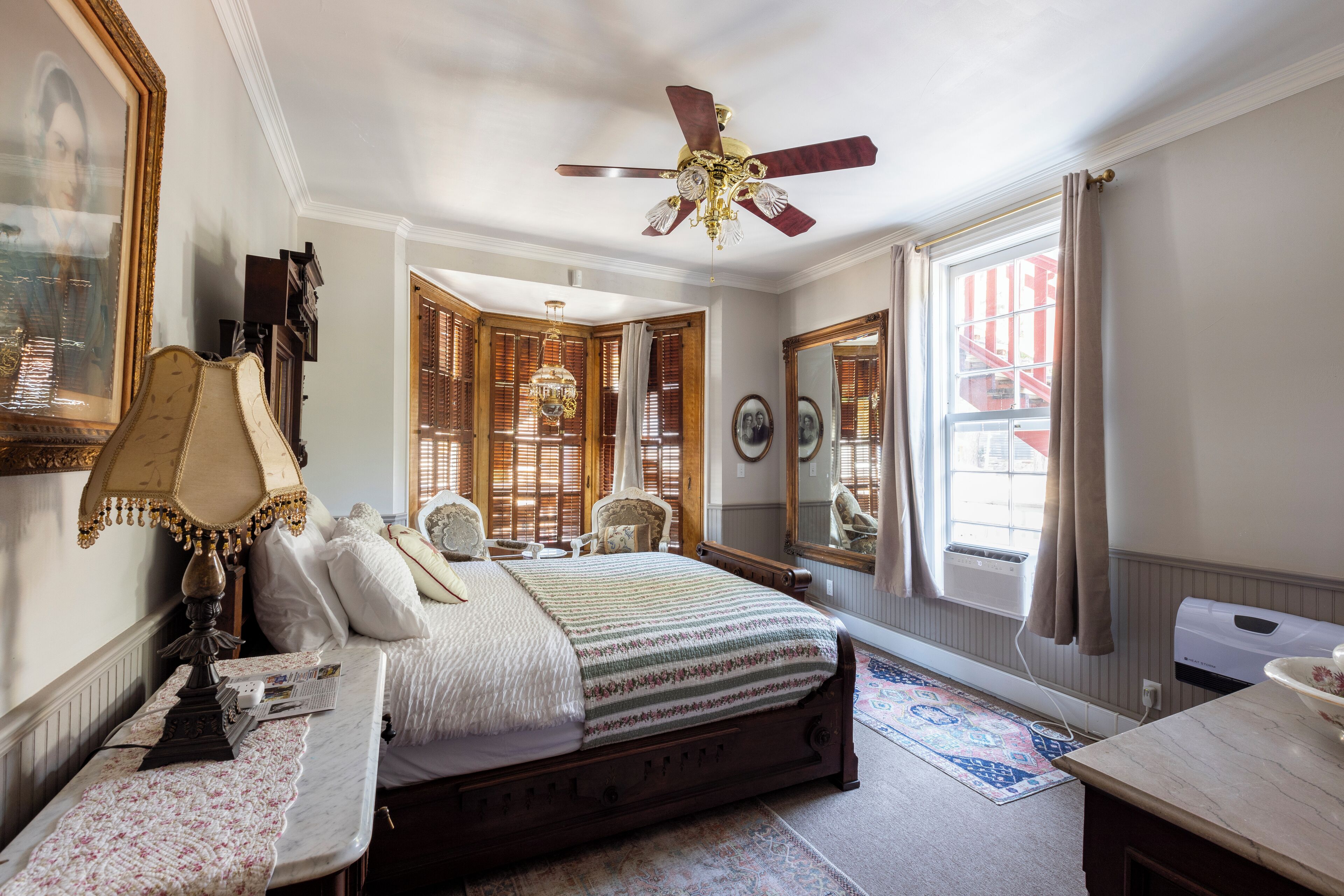 A bedroom with a large bed, a ceiling fan, a window with curtains, a mirror, and a radiator.