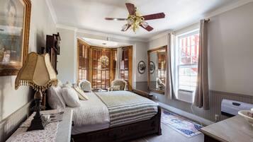 Un dormitorio con una cama grande, un ventilador de techo, una ventana con cortinas, un espejo y un radiador.