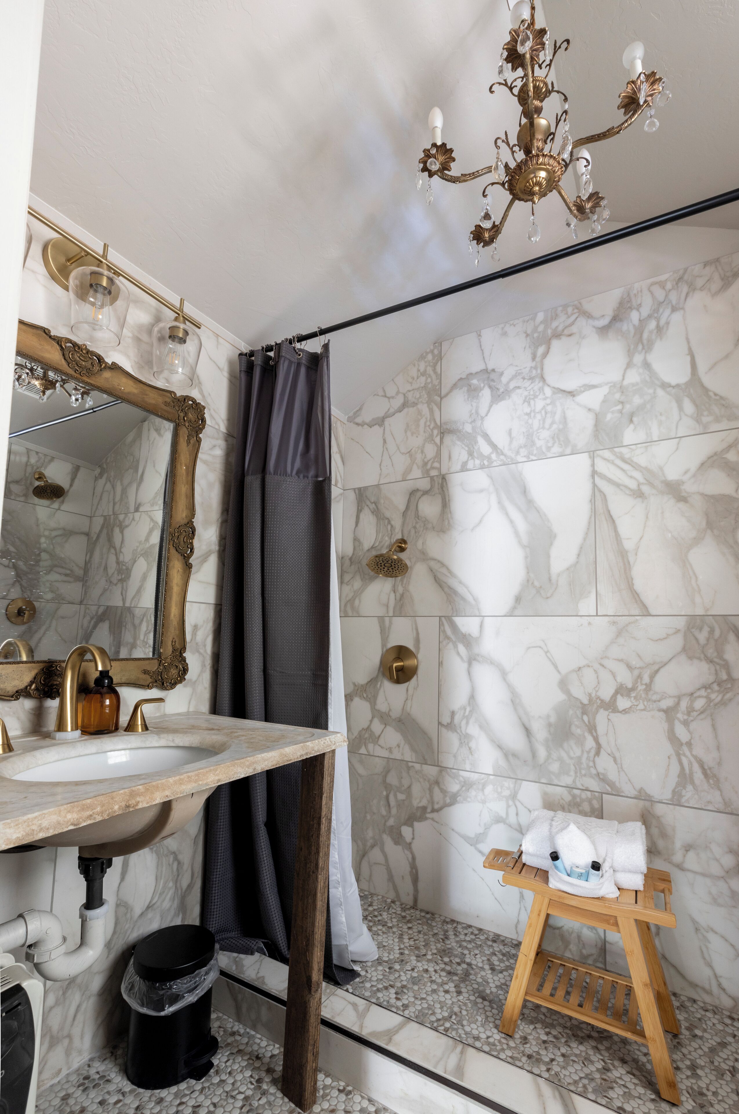Un baño con pared de mármol, espejo con marco dorado, tocador de madera, lámpara de araña y un pequeño taburete con toallas.