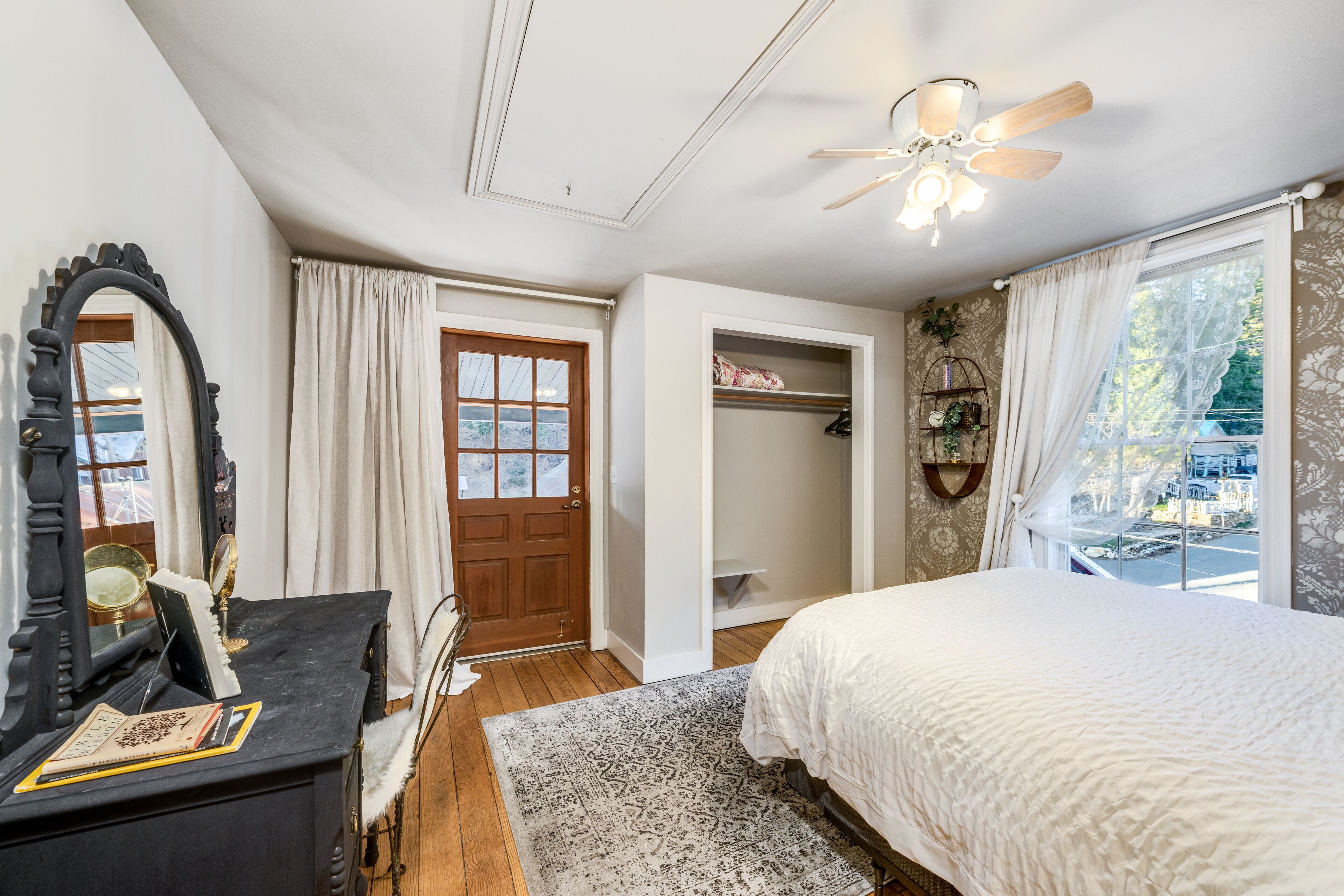 Une chambre à coucher comprenant un lit, une commode, un miroir, un ventilateur de plafond et une fenêtre avec des rideaux.
