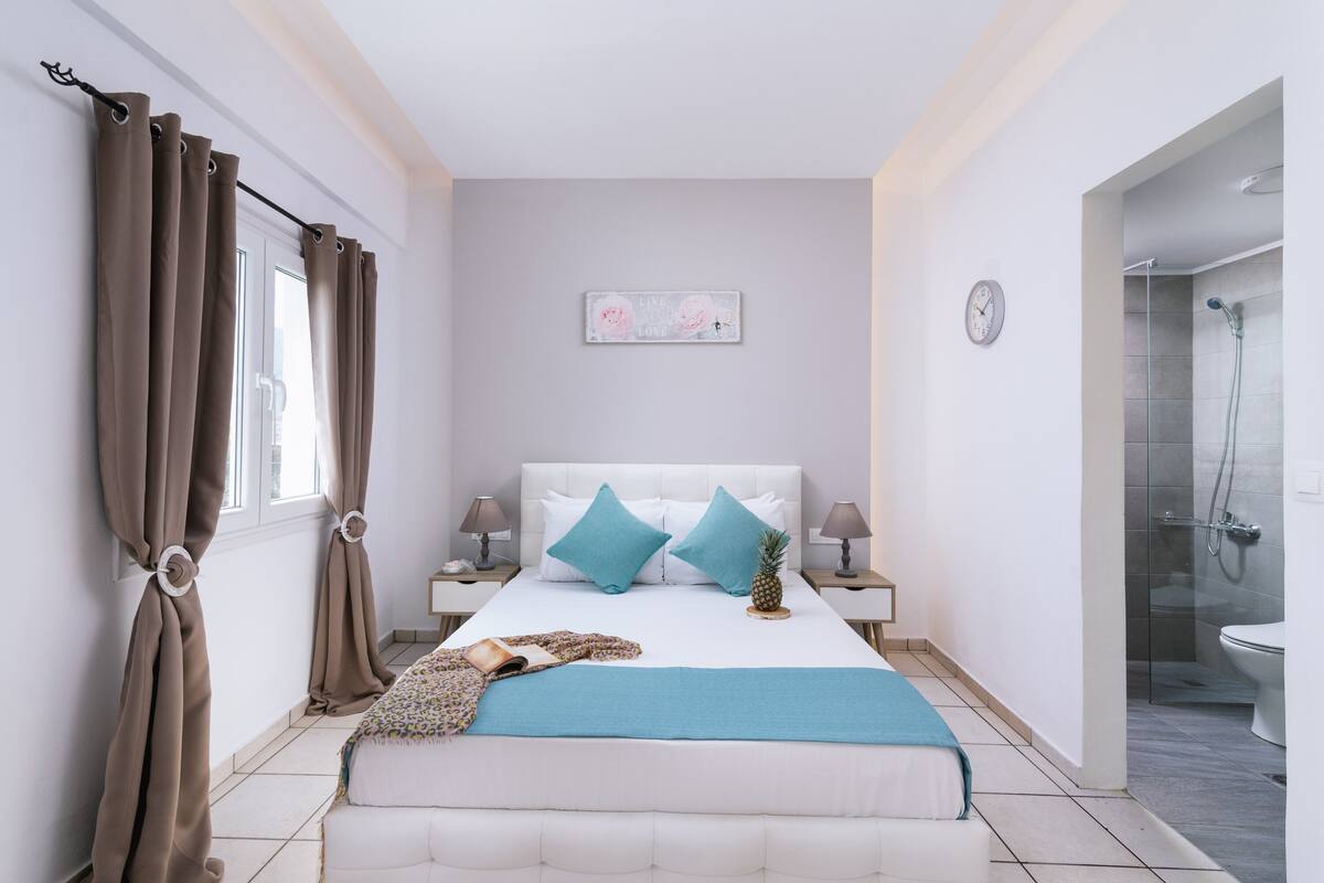 A bedroom with a bed, two nightstands, a clock, a painting, and a bathroom with a shower and toilet.