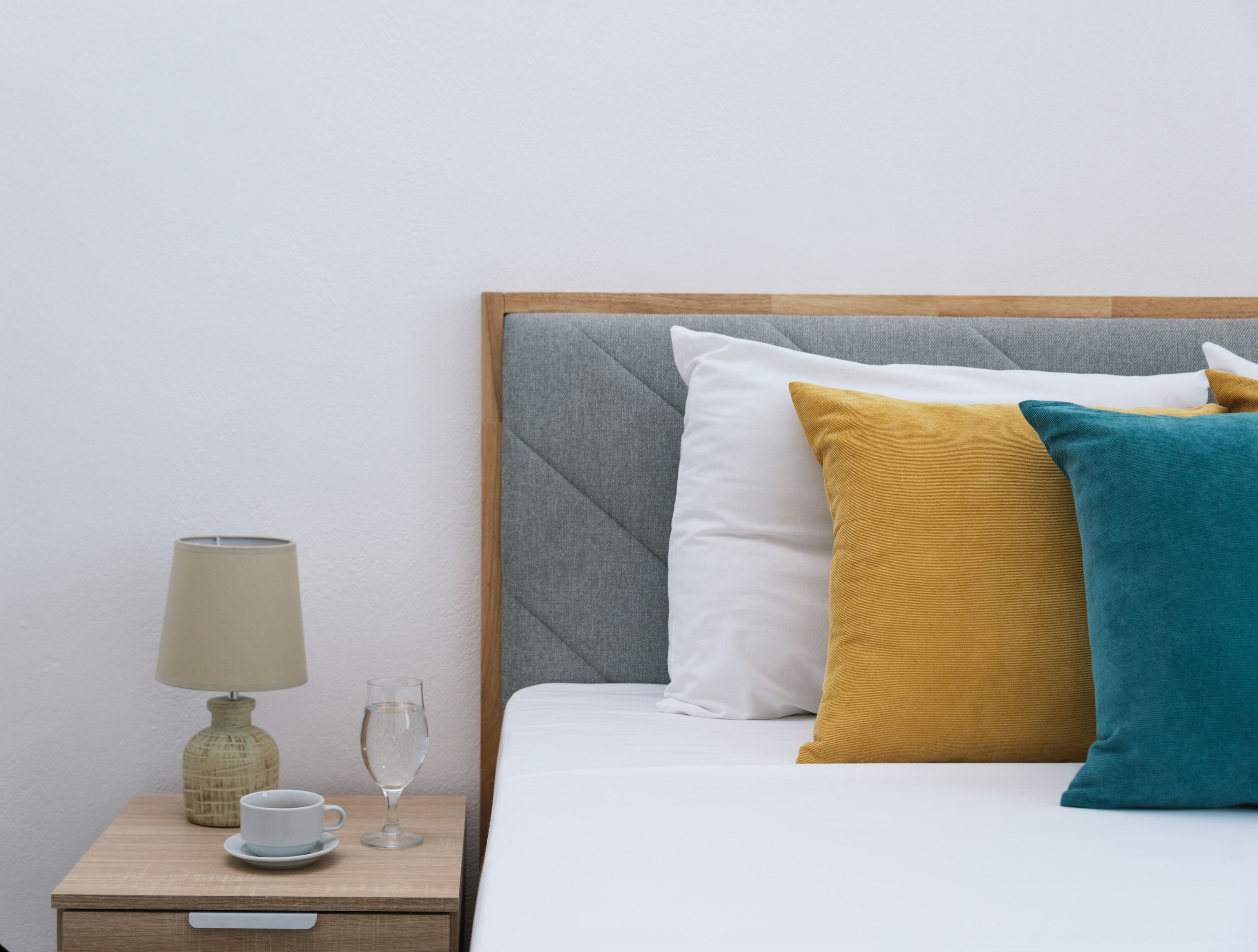A neatly made bed with a grey headboard, accompanied by a bedside table with a lamp, a cup, and a glass.