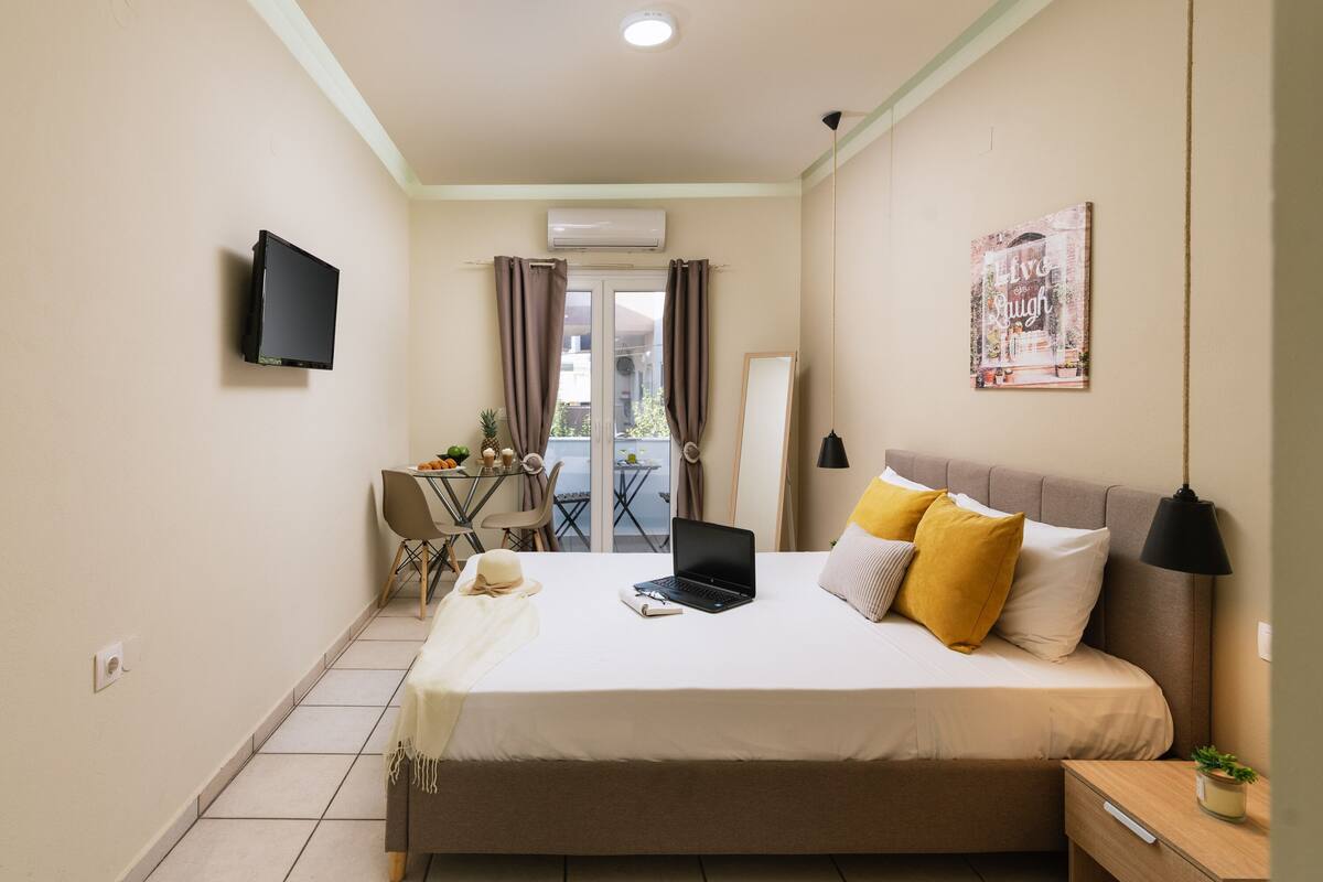 A bedroom with a bed, a desk, a chair, a laptop, a TV, and a balcony with curtains.