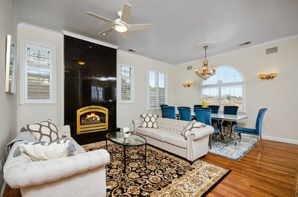 Cozy living space with a fireplace, comfortable seating, and dining area, featuring a ceiling fan and large window.