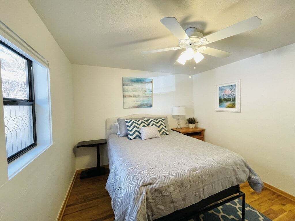 A bedroom with a bed, a ceiling fan, a window, and two wall-mounted paintings.