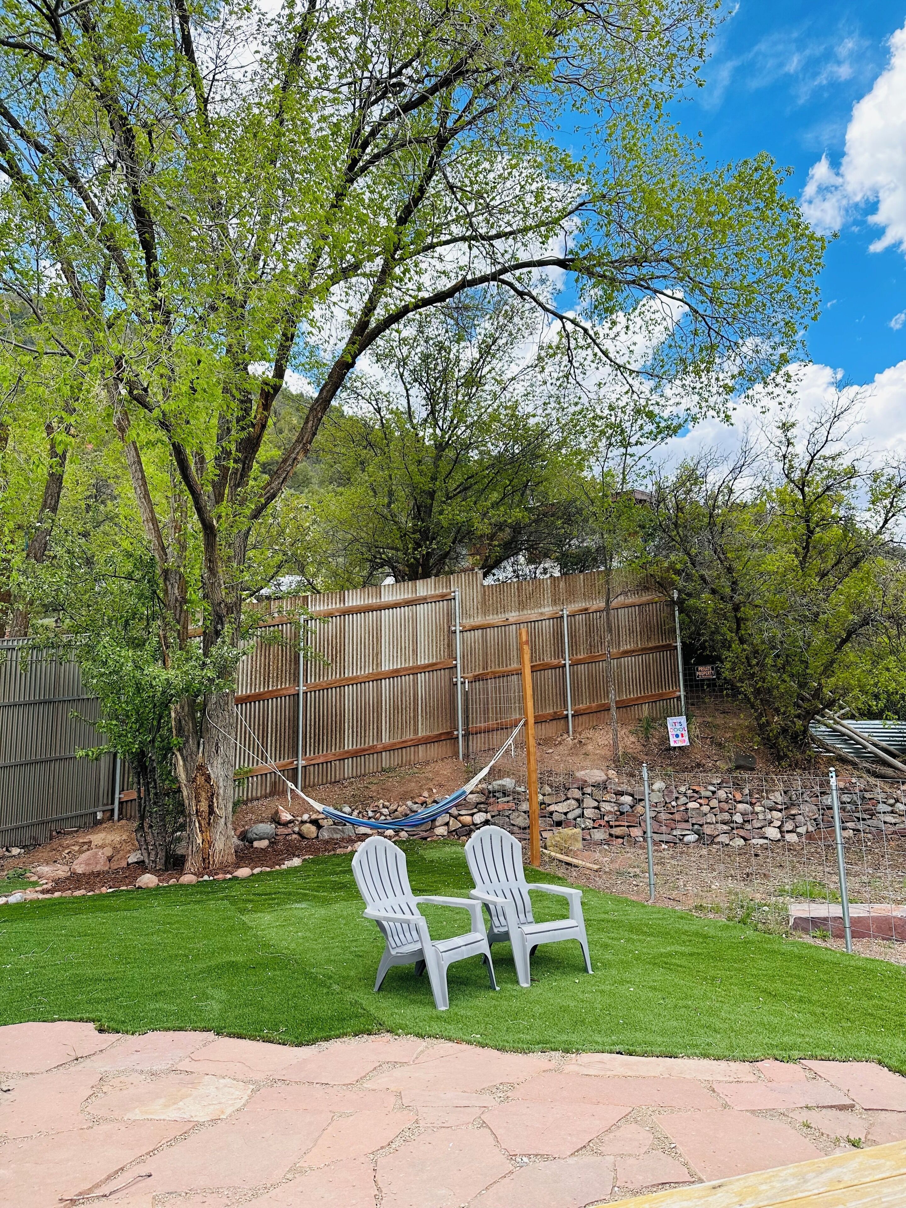 A backyard with a hammock, two Adirondack chairs, a wooden fence, and a stone wall.