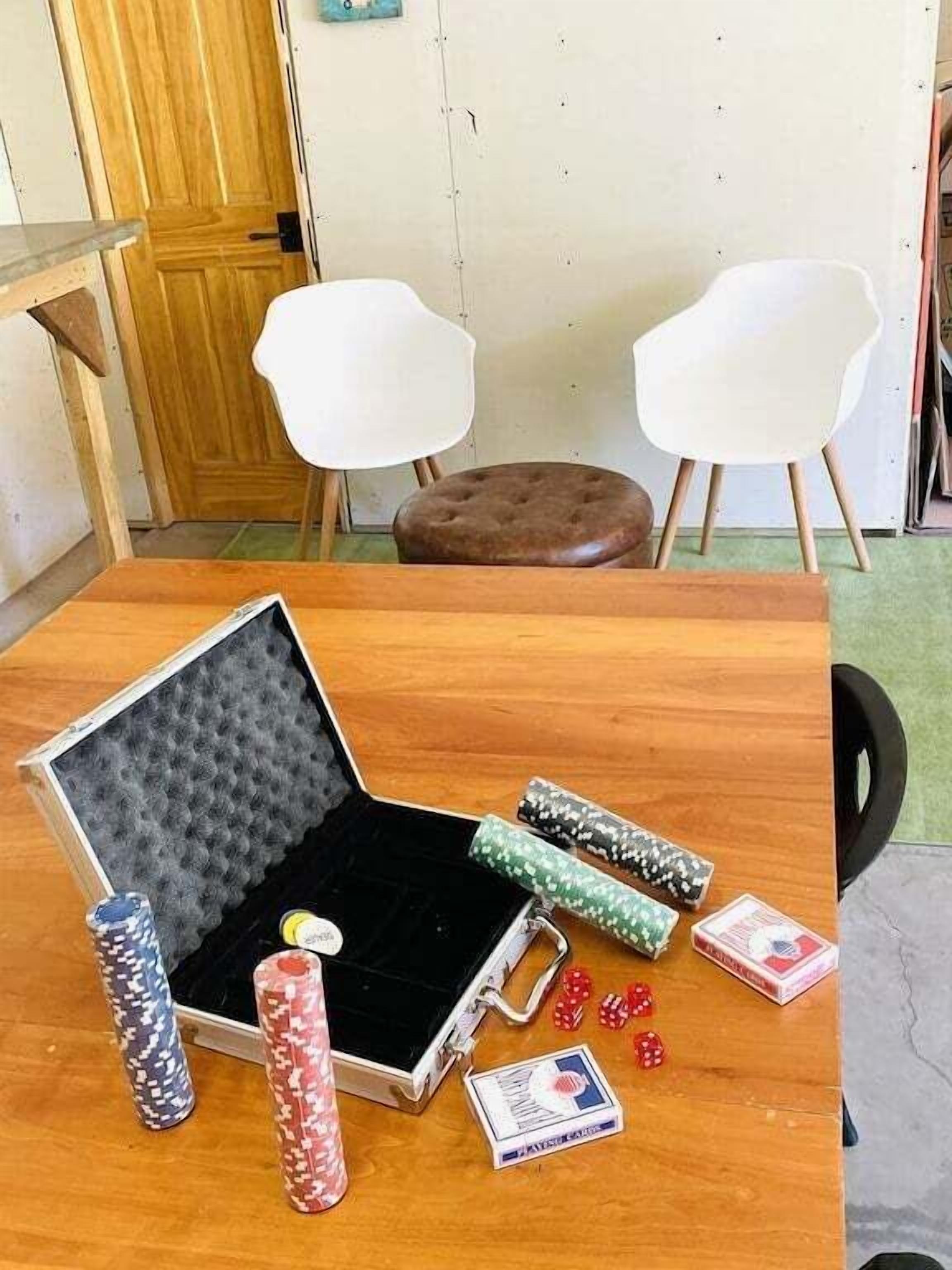 A wooden table with a poker set, including chips and playing cards, in a room with two white chairs and a brown stool.