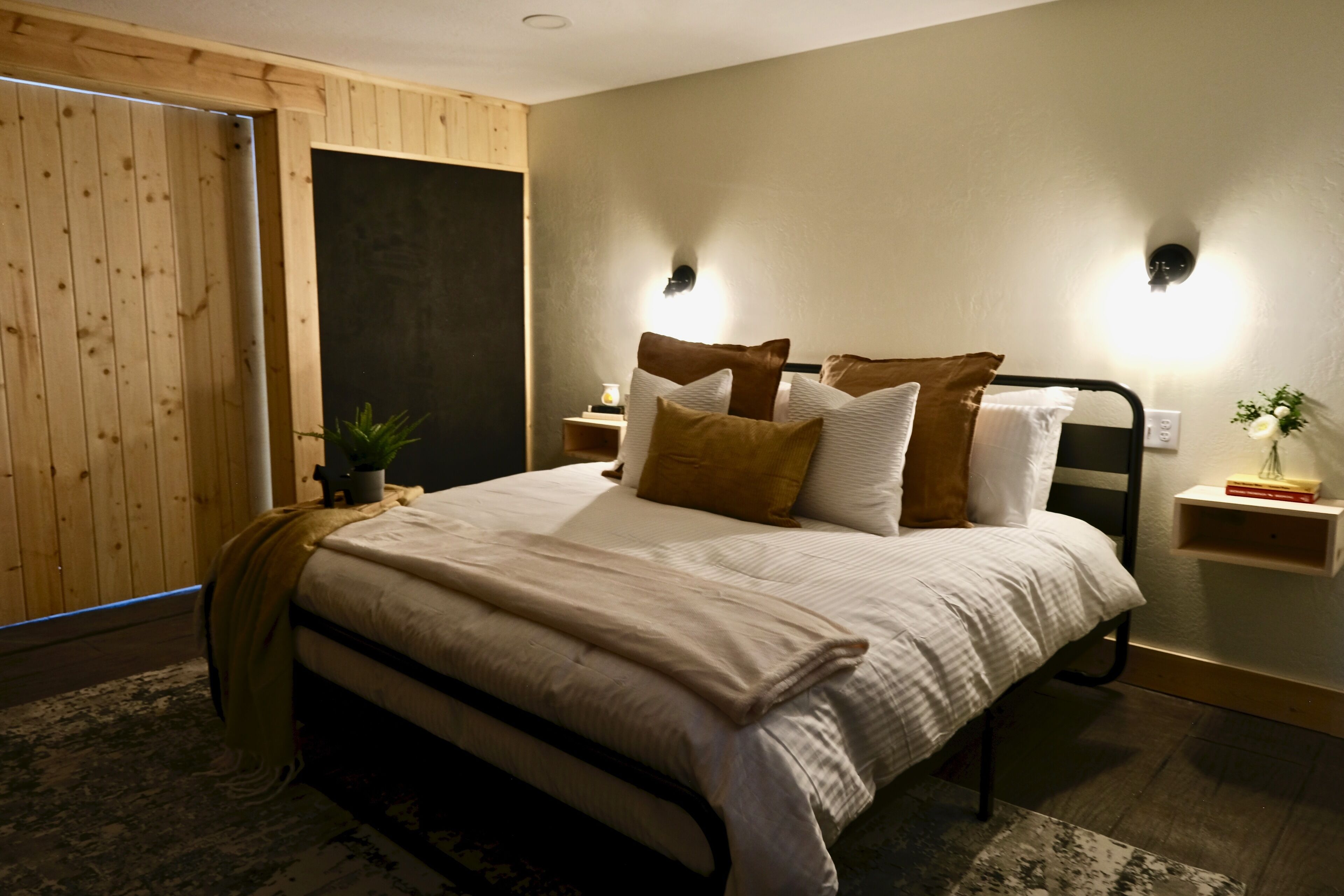A bedroom with a bed, bedside tables, a nightstand, a potted plant, and a wooden door.