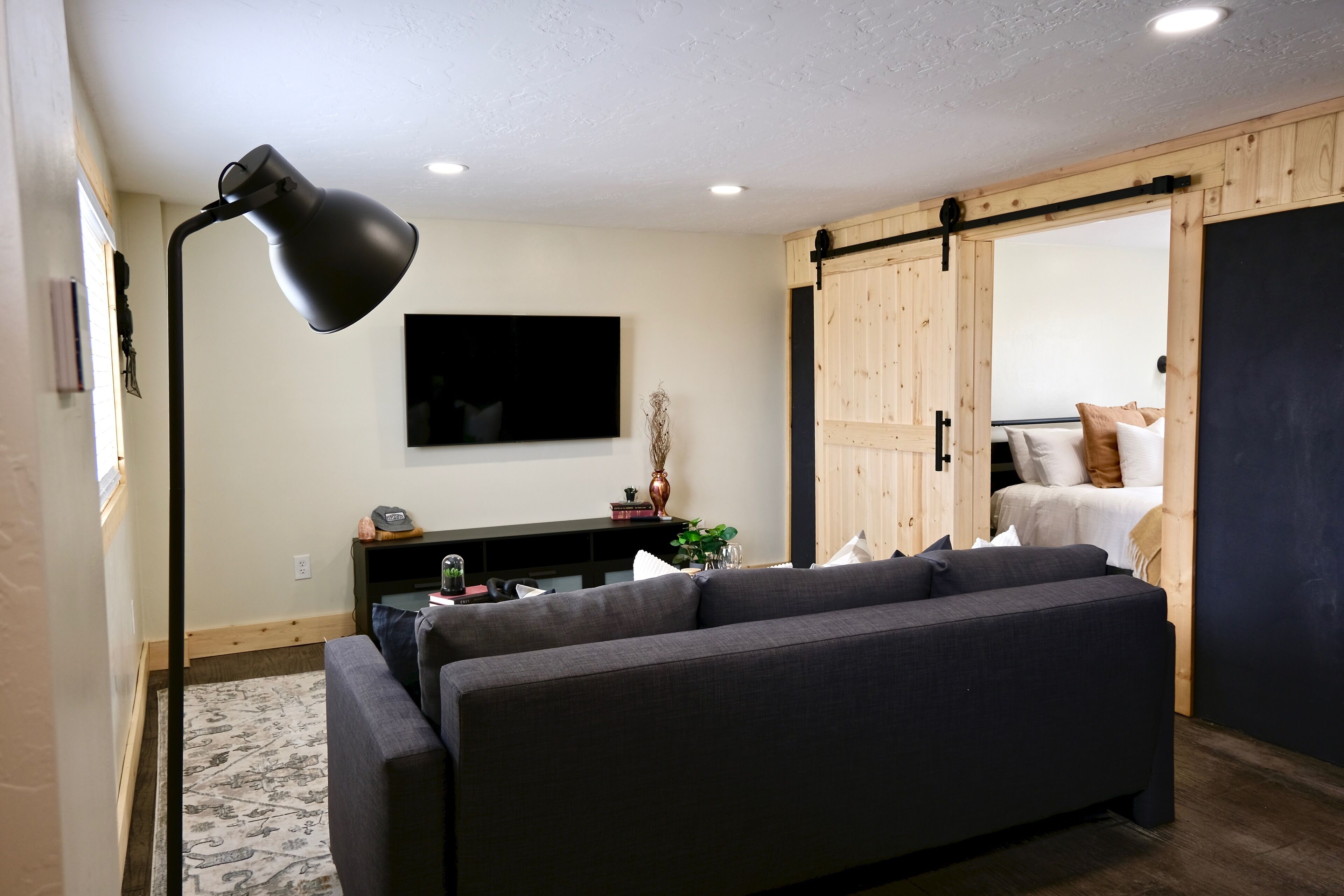 A modern living room with a dark sofa, a floor lamp, a flat-screen TV, and a sliding wooden door leading to a bedroom.
