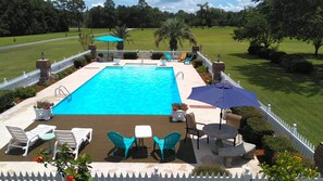 Ein rechteckiger Swimmingpool mit einer weißen Liegefläche, umgeben von einem weißen Zaun. Rings um den Pool stehen mehrere Stühle und Tische, und in der Nähe ist ein blauer Sonnenschirm aufgestellt.