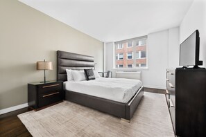 A modern bedroom with a large bed, a nightstand with a lamp, a dresser, and a television.