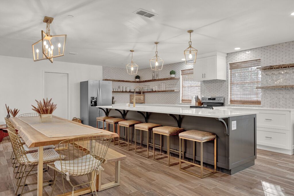 A modern kitchen with a central island, bar stools, pendant lighting, and a dining area with a long table and chairs.