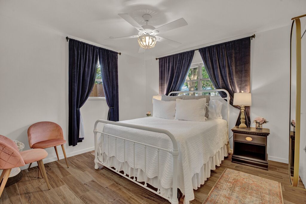 Quarto com cama branca, cortinas escuras, ventoinha no teto, mesa de cabeceira com luminária e uma cadeira.