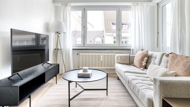 A modern living room with a flat-screen TV, a standing lamp, a round coffee table, and a sofa with patterned cushions. - Sleek Iselin 2 Room w/ Storage, 1min to Local Market, by Blueground (Basel)