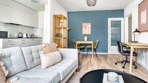A modern apartment with a grey sofa, a wooden dining table with two chairs, a small desk with a chair, and a kitchen area with white cabinets and a microwave. - Sleek Iselin 2 Room w/ Storage, 1min to Local Market, by Blueground (Basel)
